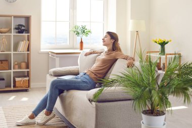 Happy relaxed woman resting on comfy couch in modern interior of her own apartment. Young girl spending free time at home, enjoying quiet leisure, sitting on cosy sofa in living room. Comfort concept