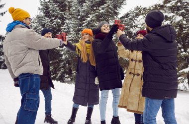 Group of cheerful friends with hot drinks in their hands warm up while walking in winter forest. Young men and women in winter clothes are clinking disposable paper cups while standing in snowy area.