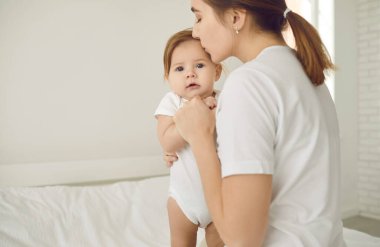 Mom together with baby. Caring young mother tenderly kissing and soothing scared child. Sweet, adorable baby girl looking at camera with expression of fear on face when she sees a stranger. Copy space