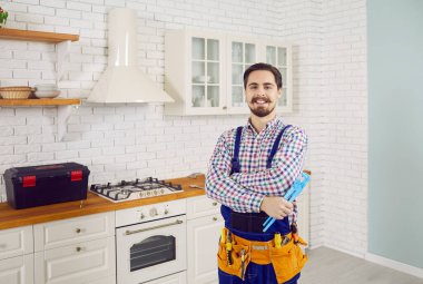 Portrait of smiling young plumber with tool in uniform pose at modern kitchen in client home. Happy mechanic or plumbing specialist repair fix water pipelines. Maintenance concept.