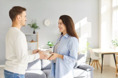 Young loving boyfriend congratulating smiling girlfriend, giving gift box in new living room after moving, woman receiving present, happy couple celebrating anniversary or birthday at home together