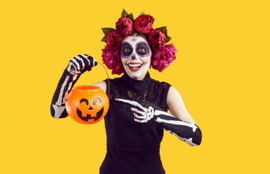 Cheerful woman in Halloween Mexican costume hints with gesture that she wants to trick or treat. Woman on orange background points with her finger to the pumpkin candy basket in her hand.