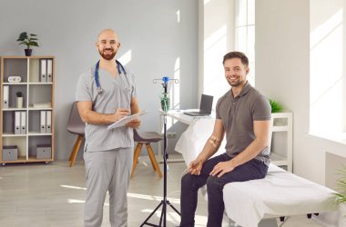 Happy male nurse together with patient. Portrait of young man sitting on medical bed and getting intravenous vitamin therapy and clinic nurse in uniform with clipboard smiling and looking at camera