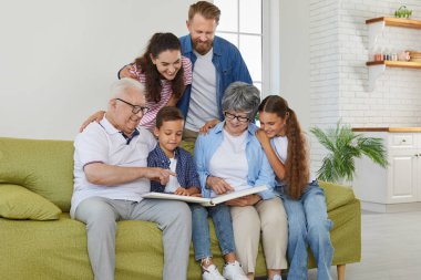 A happy family sits on a sofa and looks at a family photo album. Grandfather, grandmother, children, grandchildren spend time looking at family photos while sitting on the sofa. Happy family concept.