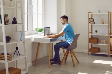 Serious focused businessman or freelancer working with laptop at home office. Young man in casual clothes sitting at desk working on project, researching, making notes on paper