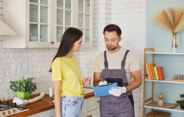 Erkek tesisatçı, kadın müşteriyle konuşuyor. Evde tesisat hizmeti için kontrat imzalıyor. Erkek tamirci ya da tamirci kadın müşteri mutfakta bekliyor. Bakım ve onarım servisi.