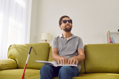 Siyah gözlüklü, sakallı, genç bir adamın portresi. Oturma odasındaki kanepede oturuyor. Sopa bastonuyla ve dokunsal kabartma kitabı okuyor. Körlük ve eğitim kavramı.