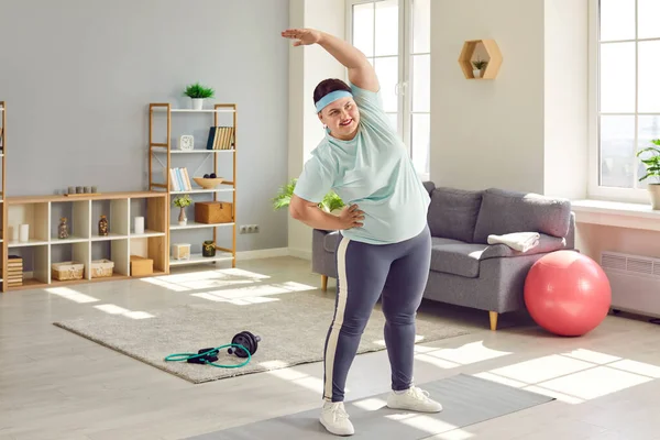 Portrait of a funny young smiling fat overweight woman wearing sportswear doing fit warming up exercise in the living room at home. Healthy lifestyle, obesity and workout sport concept.