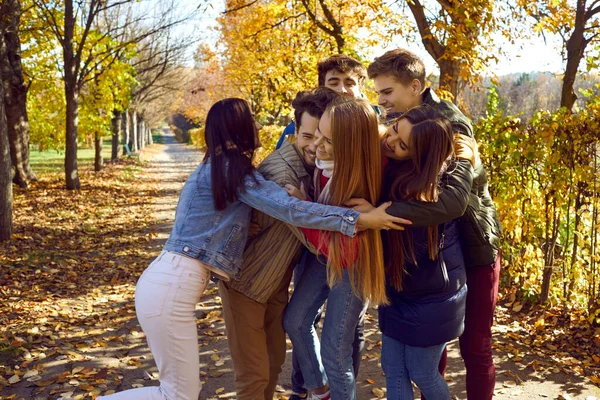 Group of happy young friends having fun in autumn park. Gathered together and hugging each other with wide smile and joyful mood against background of autumn park and yellow-orange leaves.
