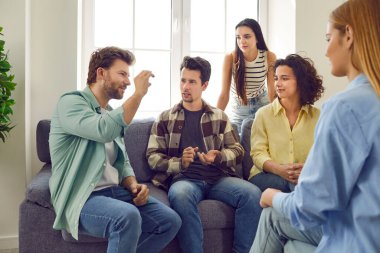 Group of friends gather at home and spend time together. Several people sitting on the couch and talking. Bunch of students playing charades. Young man tells a story or explains a word to his pals