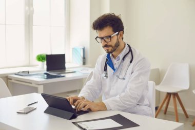 Busy young doctor using computer. Serious Caucasian male physician in medical lab coat and glasses sitting at office desk and working with electronic documents on modern digital tablet with keyboard