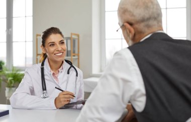 Friendly young female doctor in hospital office consults senior male patient and makes entries in his medical card. Smiling nurse sitting at table talking to mature man sitting with his back to camera