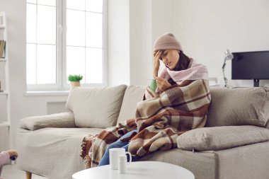 Sick woman who has fever looks frustrated at thermometer which again shows high temperature. Worried woman sitting at home on sofa wrapped in plaid, scarf and wearing hat. Concept of viral infection.
