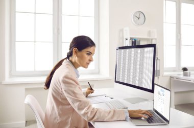 Serious businesswoman using two computers in her office. Focused young woman in suit sitting at desk, working on desktop and laptop computers, checking information and taking notes on paper