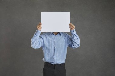 Studio portrait of unrecognizable young man covering his face hiding behind blank mockup banner. Anonymous businessman holding clean empty white sheet of paper standing isolated on grey background