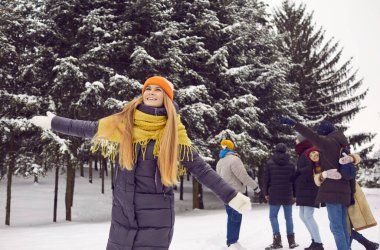 Cheerful and carefree young woman having fun in winter forest against background of her friends. Beautiful girl in winter clothes stands with arms outstretched and looks up with smile.