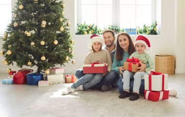 Mutlu gülümseyen anne, baba ve iki küçük çocuğun portresi Noel ağacının altında oturma odasında bir sürü hediye kutusuyla oturuyorlar. Kış tatili kavramı.
