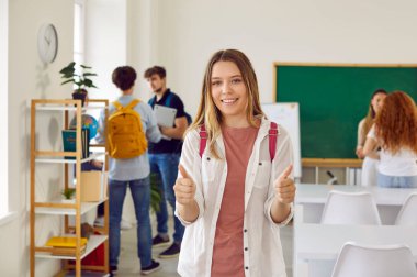 Mutlu okul, üniversite ya da üniversite öğrencisi. Güzel genç bir kadın ya da genç bir kız gülümser ve sınıfta arka planda diğer öğrencilerle birlikte başparmak kaldırır. Başarı konsepti