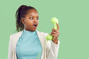 Genç siyahi kadın telefonla şok edici bir haber alıyor ve alıcıya ağzı açık ve şaşırmış bir yüz ifadesiyle bakıyor. Yeşil arkaplana karşı stüdyo portresi