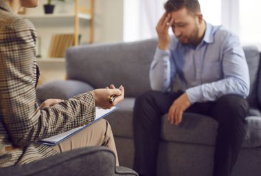 Not panosu olan kadın terapist arka planda oturan bir adamla konuşuyor üzgün, çaresiz ve kafası karışmış bir şekilde sorunlarını paylaşacak cesareti bulmaya ve yardım istemeye çalışıyor. Depresyon terapisi konsepti