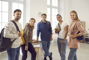 Bir grup mutlu öğrenci arkadaş. Modern sınıfta sırt çantalı, gülümseyen ve kameraya bakan beş pozitif karışık ırk ve üniversite arkadaşının kapalı alanda portresi.