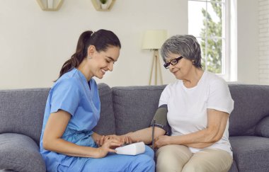 Dost canlısı doktor yüksek tansiyonlu yaşlı hastasını ziyaret ediyor. Mavi önlüklü gülümseyen tıp uzmanı. Mutlu yaşlı bir kadınla koltukta oturuyor ve kan basıncını tonometreyle ölçüyor.