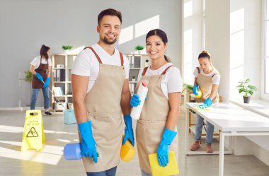Neşeli genç adam ve kadın hademeler ellerinde temizlik malzemeleri ve arka planda temizlik hizmetleri ile kameraya bakıyor. Ev işleri ve ev idaresi kavramı.
