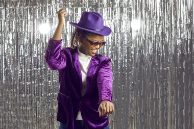 Afro-Amerikalı bir kadının disko partisinde eğlenmesi ne güzel. Mor şapkalı, kadife ceketli ve parti gözlüklü parlak gümüş folyo bir arka planda dans eden neşeli siyah kız.