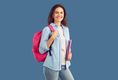Sırt çantası ve mavi arka planda fotokopi kitapları olan sırıtan esmer, çekici bir bayan öğrenci kameraya bakıyor. O kot pantolon ve mavi tişört giyiyor. Eğitim, reklam konsepti afişi.