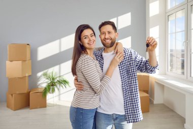 Arka planda karton kutular olan yeni bir daireden anahtar tutan mutlu genç bir çift, kucaklaşıyorlar, kameraya bakıyorlar ve kutlama günü gülümsüyorlar. Taşınma, emlak, ipotek konsepti.