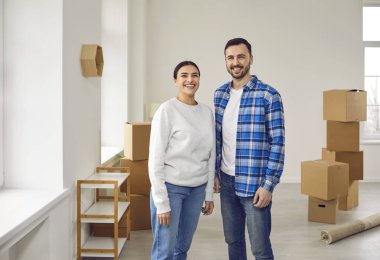 Genç çift yeni evlerine taşınıyor. Neşeli eş ev sahipleri oturma odasında karton kutularla poz veriyor ve kameraya gülümsüyor. Ev taşınması, aile konutları, gayrimenkul