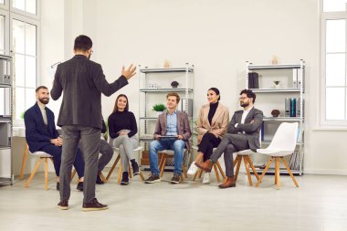 İş eğitimi. İş adamlarından oluşan küçük bir ekip, ofiste erkek konuşmacıların önünde konuşmalarını dinliyor. İnsanlar sandalyelere oturur ve önlerinde duran iş adamlarını dinlerler..