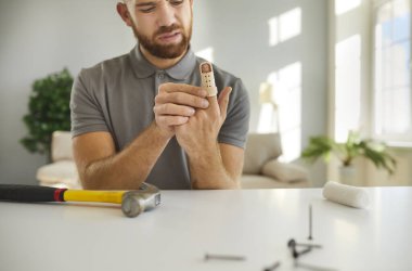 Yaralı adam evde çekiçle çalıştıktan sonra düzeltilebilir parmak bandı takar. Genç sakallı adam çekiç ve çivilerle masada oturuyor. Evde yaralanma tedavisi kavramı.