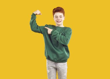 Kızıl saçlı, gülümseyen, kendine güveni tam bir genç, yeşil kazak giymiş, bir kolunda biseps olan, diğer eliyle onu işaret eden. Parlak sarı arkaplanda izole edilmiş bir stüdyo fotoğrafı..