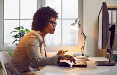 Ofiste çalışan profesyonel muhasebeci. Yan, masada oturan genç Afrikalı Amerikalı kadının profil görüntüsü, masaüstü bilgisayarının ekranına bakıyor, dijital iş sayfaları kullanıyor, verilerle çalışıyor.