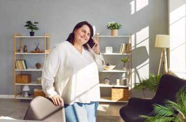 Cep telefonuyla konuşan mutlu obez kadın. Kilolu bir kız arkadaşını arıyor. Evdeki oturma odasında duran, akıllı telefonunu tutan, arkadaşını dinleyen ve gülümseyen şişman genç bayan.