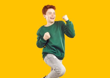 Mutlu, heyecanlı çocuğun stüdyo çekimi. Aşırı neşeli, yeşil tişörtlü, mutlu, gülümseyen yüz ifadeli, turuncu arka planda izole edilmiş yumruk hareketi yapan küçük kızıl çocuk. Başarı konsepti