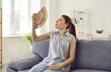 Evde sıcaktan ve yüksek sıcaklıktan muzdarip el yelpazesi kullanan stresli kadının portresi. Klimasız bir şekilde kanepede oturan hasta bir kız sıcak havadan acı çekiyor..
