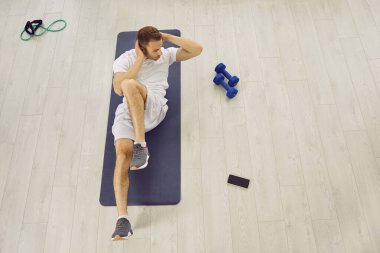 Yukarıdan bakıldığında, çapraz mekik çeken ya da halterli, dirençli bant ve cep telefonlu beyaz ahşap spor zemininde lastik spor minderi üzerinde oturan bir adam görülüyor. Spor konsepti