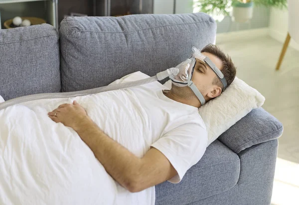 Oksijen maskesi takan adam kanepede uyuyor. Hasta evde yastık üzerinde CPAP terapisi kullanıyor. Borular bir cihaza bağlanıyor, sağlık desteği ve iyileşme öneriliyor. Konsept: Evde solunum desteği.