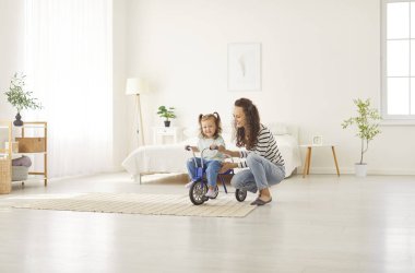Neşeli genç anne, güzel kızına geniş, parlak yatak odasında bisiklet sürmeyi öğretiyor. Küçük kız annesiyle bisiklet pratiği yapıyor, evde eğleniyor. Çocuk konseptiyle aile eğlencesi.