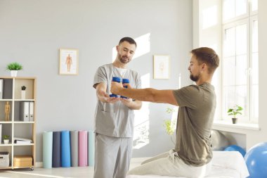 Genç adam klinikteki erkek hemşirenin desteğiyle dambıl kullanarak spor yapıyor. Fizyoterapist hastaya dambıl kaldırmada yardım ediyor. Fizyoterapi ve fizik tedavi konsepti.