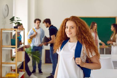 Lise, üniversite ya da üniversite eğitimi. Sınıftaki güzel, kızıl saçlı kız öğrencinin portresi sınıf arkadaşlarının arka planına karşı. Sırt çantalı kız kameraya bakıyor.