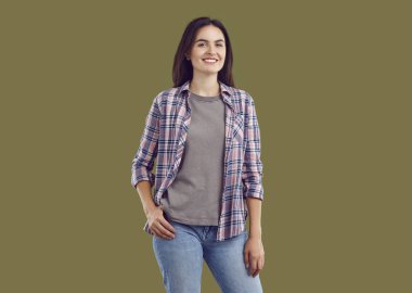 Günlük kıyafetlerle gülümseyen genç bir kadının portresi. Rahat, esmer, günlük kıyafetler giyen, elleri kalçasında, izole edilmiş haki rengi arka plandaki kameraya bakan bir kız.