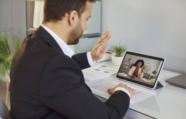 İş adamı görüntülü görüşme yapıyor. Beyaz erkek girişimci ofis masasında oturuyor, tablet ekranına el sallıyor, Afrikalı Amerikalı meslektaşını selamlıyor. Çevrimiçi iletişim kavramı