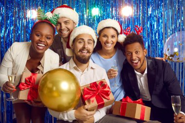 Yeni yıl partisi. Yeni yıl partisinde birlikte eğlenen neşeli, çok ırklı genç insanların portresi. Şampanya kadehleri ve hediye kutuları olan arkadaşlar parlak folyo perdelerin arkasında poz veriyorlar..