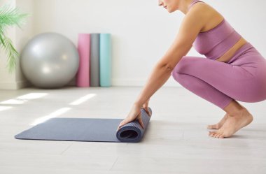 Spor giyimli formalı genç bir kadın arka planda egzersiz topuyla yoga minderini yere seriyor. Aktif kadın antrenman, esneme seansı ya da spor salonunda yoga dersi için hazırlanıyor..