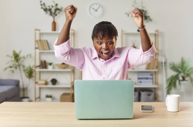 Mutlu, heyecanlı, genç, Afro-Amerikan kadın ofiste dizüstü bilgisayarda çalışıyor ve başarılı bir işte mutlu bir jest yapıyor. Başarı ve iş başarısı kavramı.