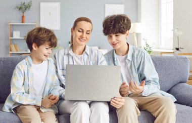 Genç anne ve oğulları video, çizgi film ya da online ders izlemek için dizüstü bilgisayar kullanıyorlar. Mutlu ebeveyn ve iki kardeş evde oturup oyun oynuyorlar, online uygulama ya da servis kullanıyorlar ve sohbet ediyorlar.