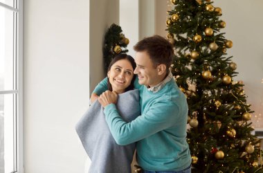 Mutlu gülümseyen genç bir çiftin portresi. Sıradan giysiler giymiş, Noel arifesinde sıcak bir battaniyeye sarılmış bir şekilde evde dikiliyor. Kış tatili kavramı.